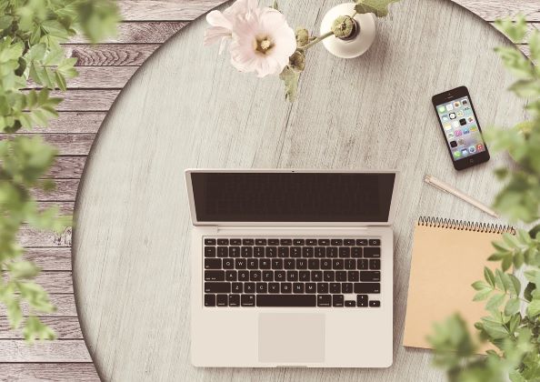 Na stoliku leżą laptop, telefon, notatnik i flakon z kwiatem.