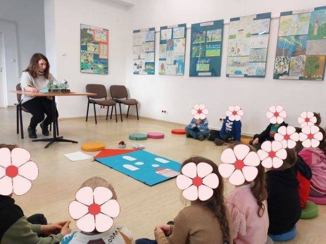 Zajęcia z dziećmi na temat przygód Muminków. Pani bibliotekarka czyta dzieciom książkę o Muminkach.Dzieci siedzą wokół domku Muminka narysowanego na dużej kartce papieru.